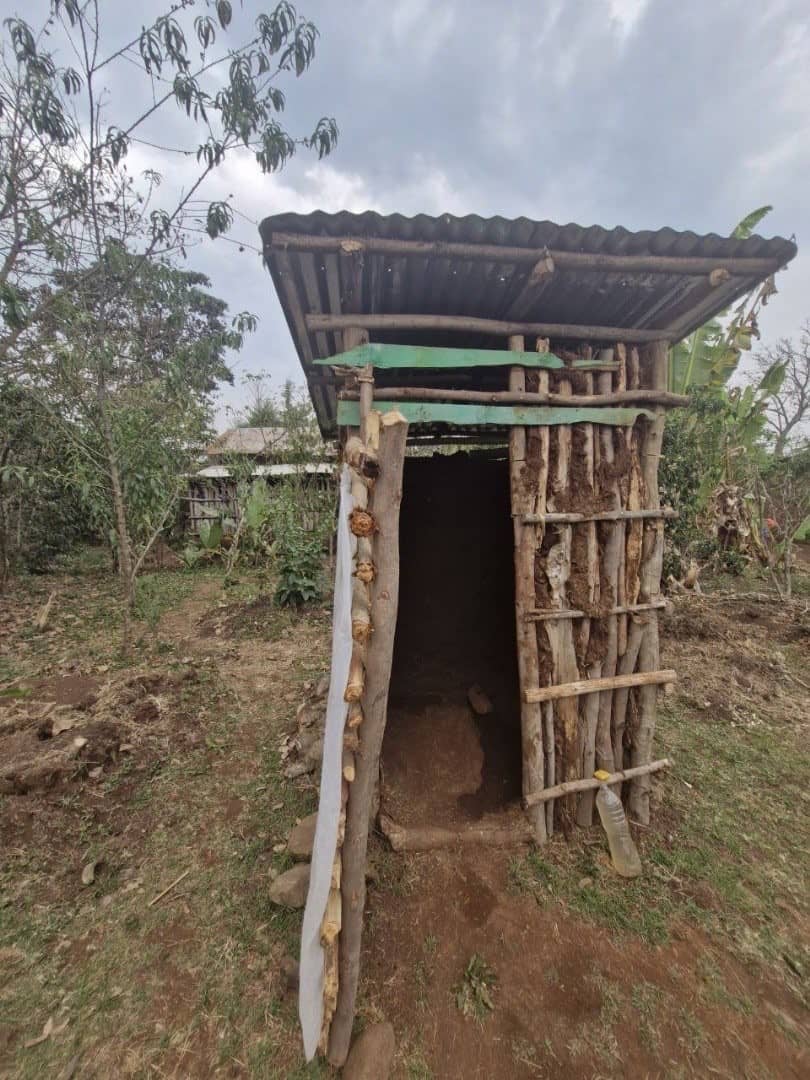 Wooden toilet cabin with corrugated iron roof. A bottle of water is placed at the entrance.