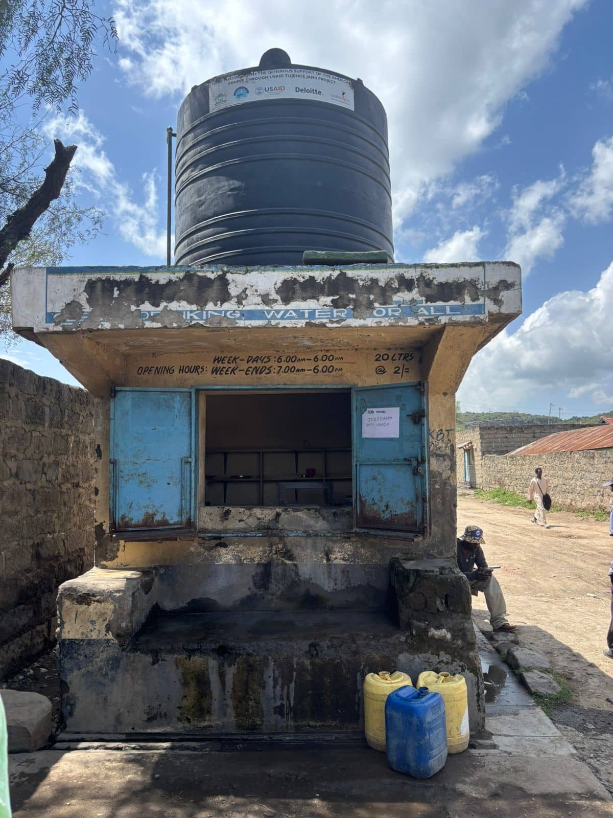 Water kiosk with water tank on roof and jerry cans in front of kiosk. Daily opening hours depicted on building.
