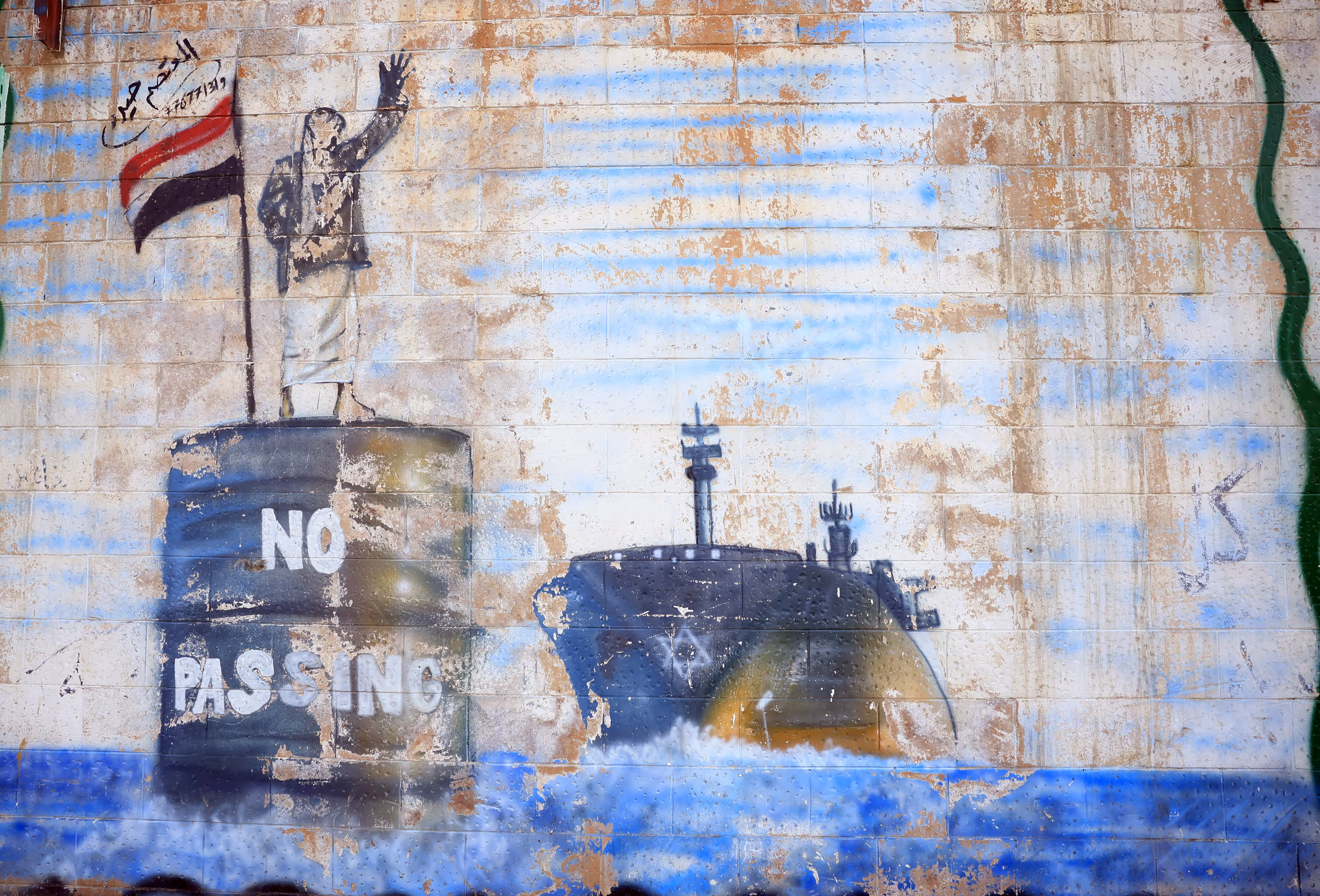 Photo of a Houthi mural. A man stands on an oil drum with the text "No Passing" next to a Yemeni flag and waves at a large ship with the Star of David on the back.